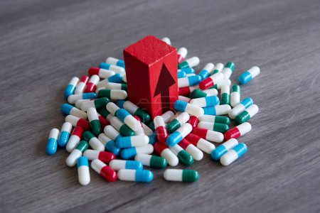 Ein roter Holzblock mit nach oben zeigendem Pfeil auf einem Stapel bunter Pillen. Steigende Kosten für verschreibungspflichtige Medikamente.