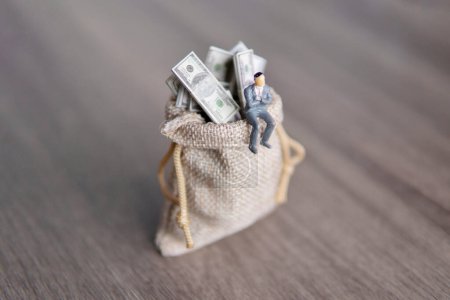 Imagen de primer plano del hombre de negocios en miniatura sentado en un gran saco desbordante de billetes de dólar estadounidense. Copia espacio para texto. Éxito, beneficio, concepto capitalista.