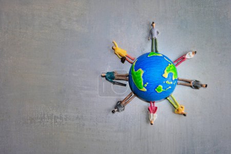 A group of miniature toy people in various colors stand around a globe on a gray surface. Copy space for text. Earth day, diversity and multiracial concept.