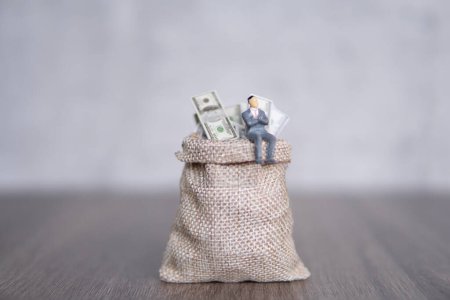 Image rapprochée d'un homme d'affaires miniature assis sur un grand sac débordant de billets en dollars américains. Espace de copie pour le texte. Succès, profit, concept capitaliste.
