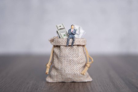 Image rapprochée d'un homme d'affaires miniature assis sur un grand sac débordant de billets en dollars américains. Espace de copie pour le texte. Succès, profit, concept capitaliste.