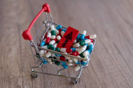 A shopping cart overflowing with colorful pills with a red arrow pointing upwards. Copy space. The rising cost of prescription drugs concept.