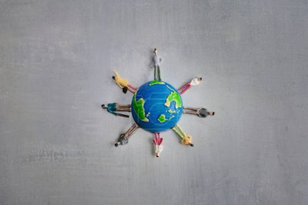 A group of miniature toy people in various colors stand around a globe on a gray surface. Copy space for text. Earth day, diversity and multiracial concept.
