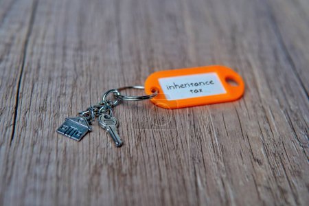A close-up photo of a keys and a house-shaped keychain on wooden table with text INHERITANCE TAX.