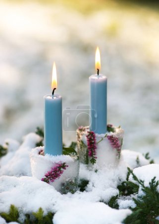 Téléchargez les photos : Beaux bougeoirs de Noël en glace, branches roses de fleur de calluna avec des bougies bleues brûlantes dans le garen recouvert de neige. Hiver extérieur concept de décoration à la main - en image libre de droit