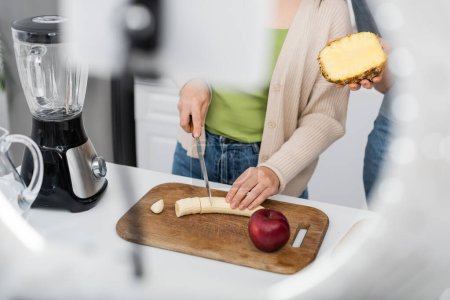 Vista recortada de las mujeres cortando plátano cerca de licuadora y teléfono inteligente borroso en la cocina 