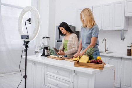 Bloggers Interraciales cortando frutas frescas cerca de licuadora y smartphone en la cocina 