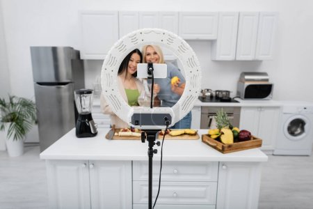 Foto de Smartphone y anillo de luz cerca borrosa amigos multiétnicos con frutas en la cocina - Imagen libre de derechos