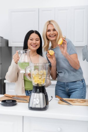 Foto de Amigos interracial positivos sosteniendo frutas y agua cerca de la licuadora en la cocina - Imagen libre de derechos