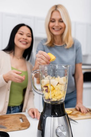 Téléchargez les photos : Blurred interracial femmes faisant smoothie aux fruits dans la cuisine - en image libre de droit