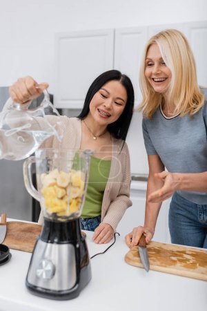 Asian woman pouring water in blender with fruits near friend in kitchen 