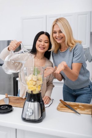 Mujer sonriente señalando con las manos cerca de amigo asiático vertiendo agua en licuadora con frutas en la cocina 