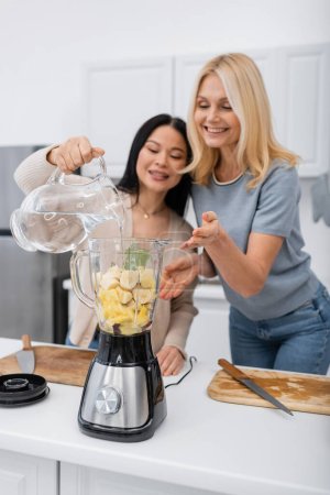 Blurred asian woman pouring water in blender while making smoothie beside friend in kitchen 