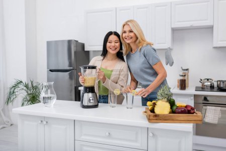 Smiling interracial women holding blender with fruit smoothie near glasses in kitchen 
