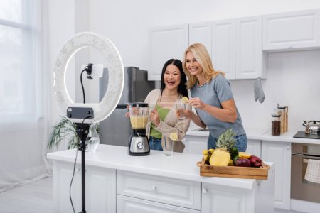 Fröhliche multiethnische Frauen halten Smoothie und Glas neben Smartphone mit Ringlampe in der Küche 