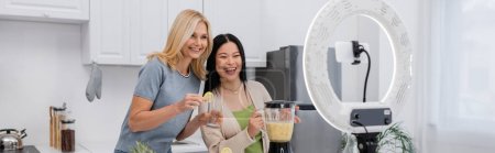 Multiethnic bloggers holding glass and blender with smoothie near smartphone and ring light at home, banner 