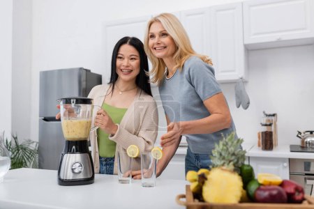 Cheerful interracial women looking away near smoothie and glasses in kitchen 