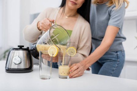 Ausgeschnittene Ansicht von Frau, die Smoothie in Glas mit Zitrone gießt, in der Nähe verschwommener Freund zu Hause 