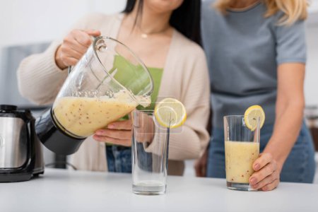 Verschwommener Blick auf verschwommene Frauen, die zu Hause Frucht-Smoothie in Gläser mit Zitronen gießen 
