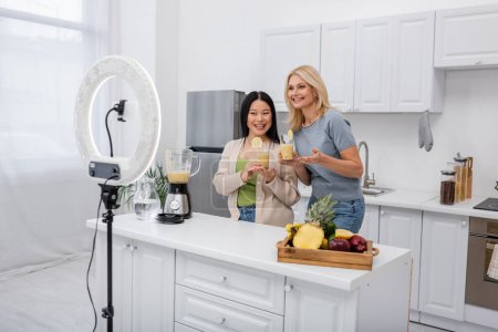 Interracial bloggers holding glasses of smoothie near cellphone and ring light in kitchen 