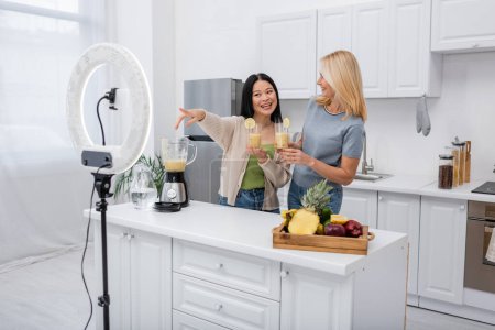 Smiling asian blogger pointing at smartphone and ring light near friend with smoothie in kitchen 