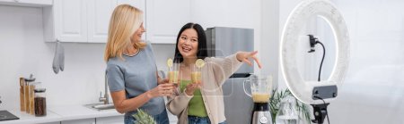Cheerful asian blogger holding smoothie and pointing at smartphone in ring light near friend at home, banner 