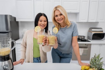 Smiling interracial friends looking at camera and holding glasses of fruit smoothie in kitchen 