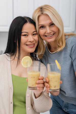 Cheerful interracial friends holding blurred glasses of smoothie at home 