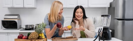 Smiling asian woman pointing at fruits near friend and smartphone on tripod in kitchen, banner 