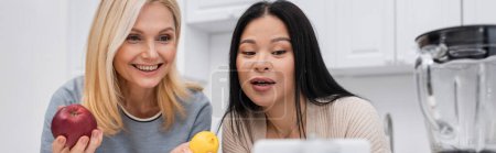 Foto de Bloggers multiétnicos sonrientes sosteniendo frutas cerca de teléfonos inteligentes en la cocina, pancarta - Imagen libre de derechos