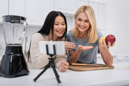 Téléchargez les photos : Femmes interraciales positives pointant du doigt les fruits près du mélangeur et du smartphone sur le trépied dans la cuisine - en image libre de droit