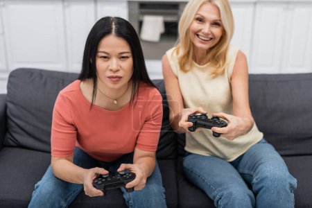 Photo for KYIV, UKRAINE - DECEMBER 2, 2021: Focused asian woman playing video game near blurred friend at home - Royalty Free Image