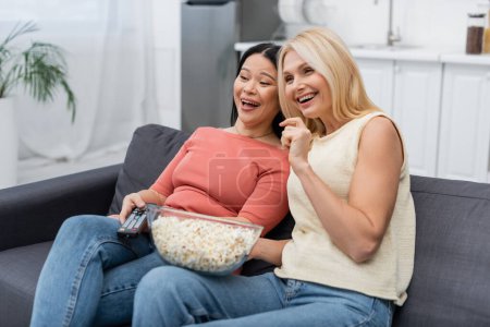 Mujeres alegres multiétnicas con mando a distancia y palomitas de maíz viendo la televisión en casa 