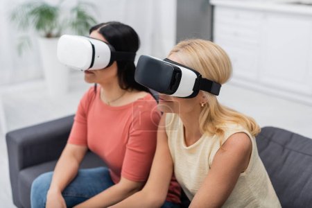 Photo for Smiling middle aged woman in vr headset gaming near blurred friend at home - Royalty Free Image