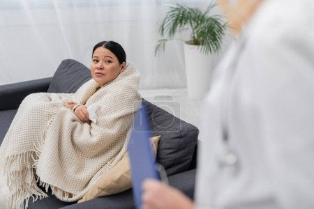 Sick asian patient in blanket looking at blurred doctor at home 