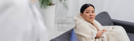 Ill asian patient holding napkin and looking at blurred doctor at home, banner 