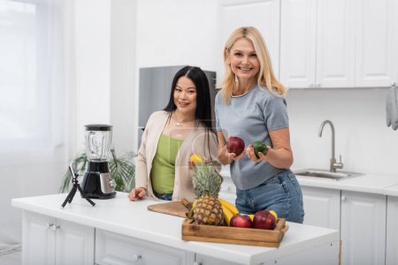 Cheerful multiethnic bloggers holding fresh fruits and looking at camera near smartphone and blender in kitchen 