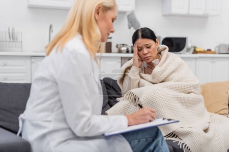 Diseased asian woman in blanket touching head near blurred doctor writing on clipboard at home 