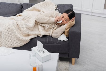 Diseased asian woman holding napkin near blurred water and pills at home 