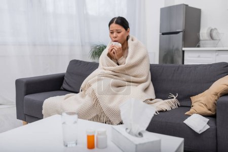 Tired and sick asian woman holding napkin near blurred pills at home 