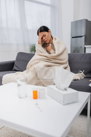 Épuisé asiatique femme dans couverture assis près de pilules et serviettes à la maison 