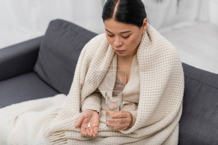 Photo for Sick asian woman in blanket holding glass of water and pills at home - Royalty Free Image