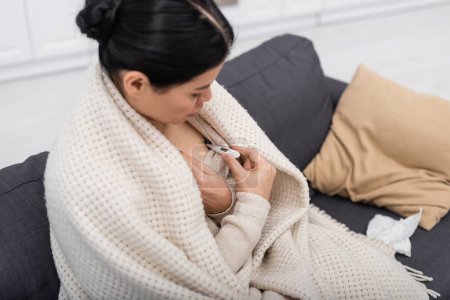 Kranke Asiatin in Decke schaut zu Hause auf elektronisches Thermometer 