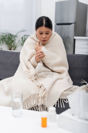 Besorgte Asiatin in Decke blickt zu Hause auf Thermometer neben verschwommenen Pillen und Servietten 
