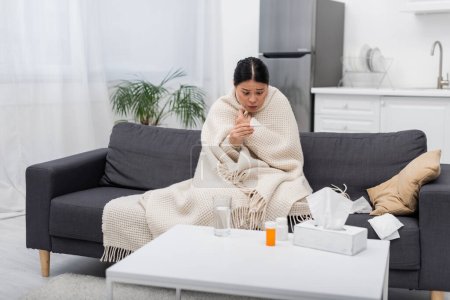 Scared asian woman in blanket looking at thermometer near pills and napkins at home 