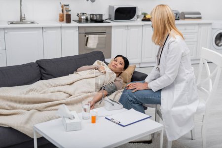 Sick asian patient lying near doctor in medical mask and tonometer at home 