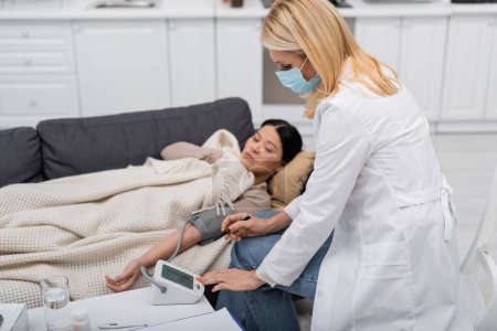 Téléchargez les photos : Docteur en masque médical regardant tonomètre près flou asiatique patient à la maison - en image libre de droit