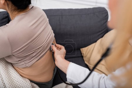 Téléchargez les photos : Vue recadrée d'un médecin flou avec stéthoscope vérifiant le dos de la femme à la maison - en image libre de droit