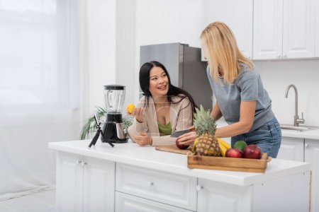 Smiling asian woman holding lemon and talking to friend cutting apple near smartphone on tripod in kitchen 