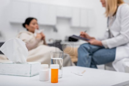 Pills and glass of water near blurred doctor and patient at home 